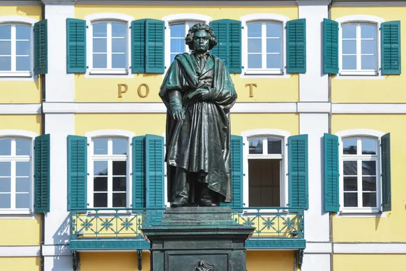 Eine Bronzestatue eines Mannes in historischer Kleidung steht vor einem gelben Gebäude mit weißen Fenstern und grünen Fensterläden. Hinter der Statue ist teilweise das Wort „POST“ sichtbar.